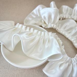 May include: Close-up of several white satin scrunchies and a small white dish. The scrunchies have a ruffled edge and are made of a shiny, smooth fabric. The items are arranged on a neutral-colored surface.