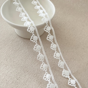 May include: Close-up of white floral lace trim. The lace features a repeating pattern of small, open flower shapes connected by a thin, woven thread. The trim is displayed against a neutral background, highlighting its delicate design.