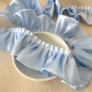 May include: Light blue satin ruffle trim, gathered and sewn, displayed over a white bowl. The fabric has a slight sheen, and the ruffles create a soft, textured effect. The trim is likely used for sewing or crafting projects.