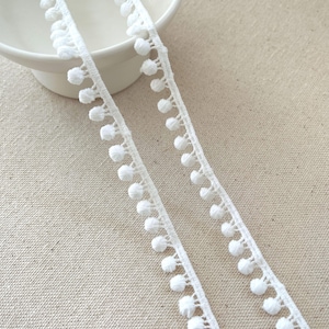 May include: Close-up of white pom-pom trim. Two strips of white trim with small, evenly spaced pom-poms are displayed against a neutral background. The trim is likely used for sewing, crafting, or adding decorative detail to fabric projects.