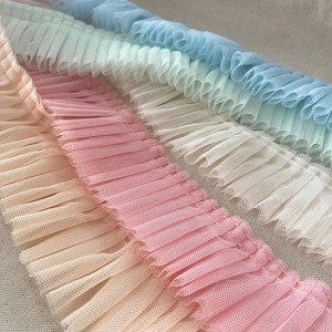 May include: Close-up of several rows of ruffled trim in pastel colors. The trim is made of a sheer, net-like fabric and features a delicate, pleated design. Colors include light peach, pink, white, light green, and light blue.