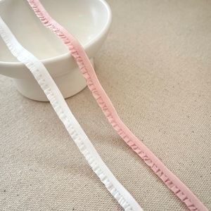 May include: Two strips of ruffled elastic ribbon in white and light pink. The ribbons are positioned diagonally across a neutral-toned surface, with a white ceramic bowl in the background. The elastic appears to be new and unused.