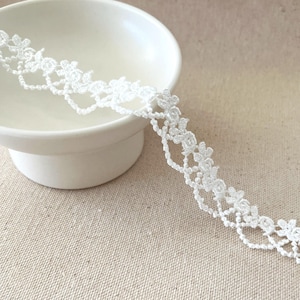 May include: White floral lace trim with a scalloped edge. The delicate lace is displayed against a neutral background and a white bowl. This trim is suitable for various craft and sewing projects.