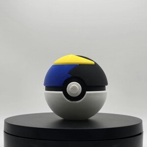 Puede incluir: Un juguete Poke Ball blanco, negro, azul y amarillo. La Poke Ball est&aacute; en una plataforma circular negra.