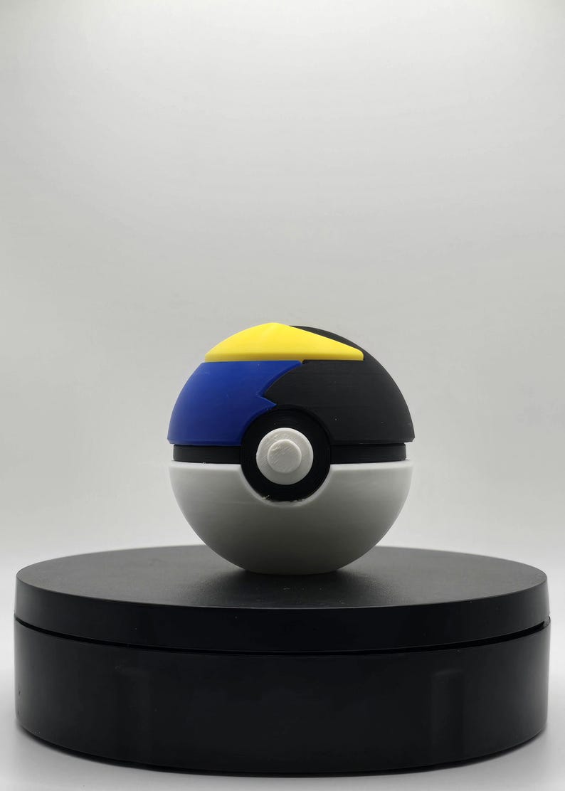 Puede incluir: Un juguete Poke Ball blanco, negro, azul y amarillo. La Poke Ball est&aacute; en una plataforma circular negra.