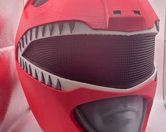 May include: A red Power Ranger helmet with a black mesh mouth and white teeth. The helmet has a gray visor and is designed with angular features. The helmet is a cosplay item.