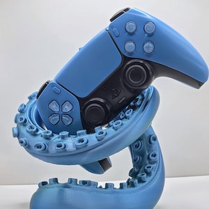 May include: A light blue octopus-shaped stand holding a blue and black video game controller. The stand is designed to cradle the controller, with the tentacles wrapping around it. The controller has buttons and directional pads.