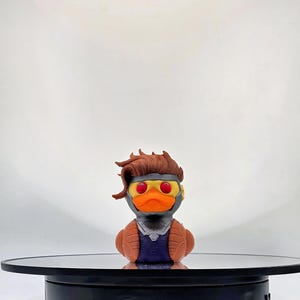 May include: A 3D-printed duck figurine with a brown hairstyle, orange beak, and yellow-rimmed goggles. The duck is wearing a brown jacket over a blue shirt. The figurine is displayed on a black rotating stand.