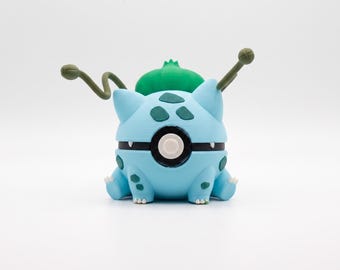 May include: A light blue spherical figurine with dark blue spots, featuring a green bulb-like structure on its back, two thin green vine-like appendages extending from the sides, and a central black and white circular button on the front.