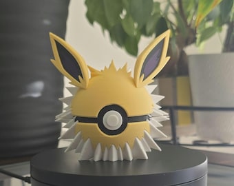May include: A yellow and white 3D printed figurine of a Pokemon character, Eevee, in its Poké Ball form. The figurine has black ears and a black Poké Ball design on its chest.