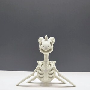 May include: A white, stylized skeleton of a cartoon creature, possibly a dinosaur or mythical beast. The figure has a long neck, rib cage, and outstretched limbs, with a smiling skull-like head. The background is a solid gray.