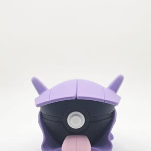 May include: A purple and black cartoon figure with a pink tongue. The figure has a rounded body with a purple head and two pointed ears. A white circle is in the center of the black body. The figure is a collectible toy.
