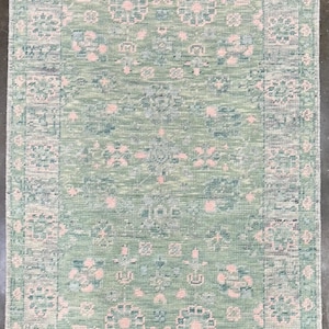 May include: Rectangular rug with a faded, vintage look. The rug features a light green background with a repeating floral pattern in pink and blue. The border has a similar design. The rug has a fringed edge.