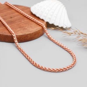 Puede incluir: Collar de cadena de color oro rosa con un diseño tejido. El collar se exhibe sobre una superficie de madera con una concha blanca y plantas secas en el fondo. La cadena es de un color metálico cálido.
