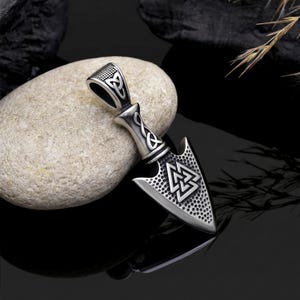 May include: Silver arrowhead pendant with Celtic knot and Valknut symbols. The pendant has a textured surface and a loop for a chain. The design is detailed and the metal has a polished finish. The pendant is likely made of stainless steel.