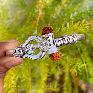 Puede incluir: Pulsera plateada con símbolos religiosos y texto. Presenta un diseño central con una gema transparente y redonda y dos cuentas marrones texturizadas. El fondo es un follaje verde borroso.