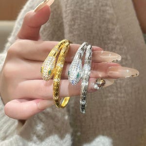 May include: Two snake-shaped bracelets, one in gold-tone metal and one in silver-tone metal. Both bracelets feature a textured scale pattern, clear pavé-set crystals covering the head and body, and small green stones serving as the snake eyes.