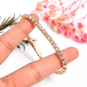 Puede incluir: Una pulsera dorada con una fila de cristales claros y brillantes. La pulsera se muestra sobre un fondo de follaje rosa y verde. Los cristales están espaciados uniformemente y capturan la luz, creando un efecto deslumbrante.