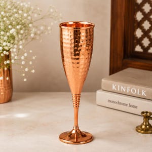 Puede incluir: Una flauta de champán de color cobre con una textura martillada. El tallo tiene un diseño acanalado y la base es redonda. La copa se exhibe sobre una superficie de color claro, con un jarrón de flores blancas y libros en el fondo.