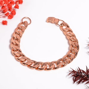 May include: A rose gold dog collar with a curb chain design. The collar features a sturdy clasp and a ring for attaching a leash. The collar is displayed on a white surface with red and burgundy floral accents.