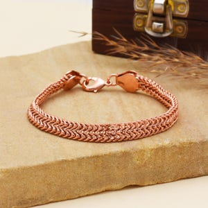 May include: A copper-colored bracelet with a woven chain design. The bracelet has a lobster clasp closure and is displayed on a light brown surface. The bracelet is approximately 7 inches in length.