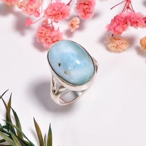 Può includere: Anello d'argento con una grande pietra ovale azzurra con inclusioni scure. L'anello è posto su uno sfondo bianco con fiori rosa e fogliame verde.