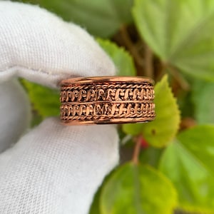 May include: A copper-colored ring with a detailed, woven design. The ring features two bands of intricate patterns separated by a simple rope-like border. The ring is held against a backdrop of green leaves.