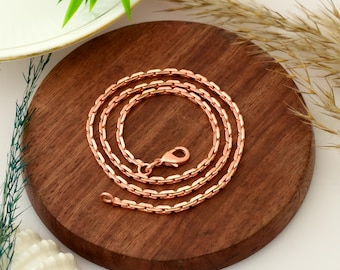 May include: A copper-colored chain necklace with a lobster clasp, coiled on a round, dark wooden surface. The necklace is made of rectangular links. The background includes a white surface, a decorative plant, and dried ornamental grass.