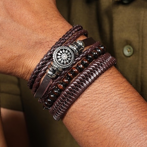 May include: A multi-layered bracelet set in shades of brown. The bracelets include braided leather, wooden beads, and a silver-toned centerpiece with a circular design. The bracelets are stacked together for a layered look.