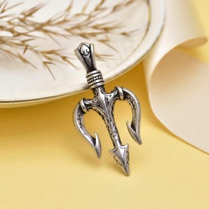 May include: A silver trident pendant with detailed patterns. The three prongs have curved ends and a central spearhead. The pendant is shown on a yellow surface, with a white plate and ribbon in the background.