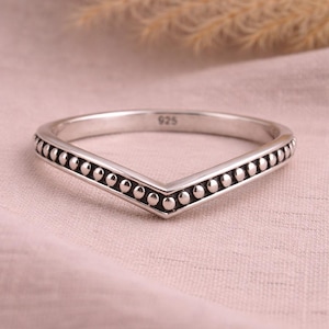 Peut inclure: Une bague argentée en forme de V avec un motif de points sur les bords. La bague est marquée "925" et est posée sur un fond rose clair. Il s'agit d'un bijou.