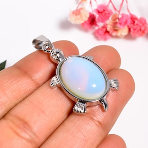Puede incluir: Colgante de tortuga plateado con una piedra ópalo ovalada. El colgante tiene una anilla para una cadena y detalles de aletas y cabeza de tortuga. La piedra tiene un suave tono blanco y azul perlado.