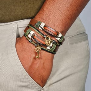Op de afbeelding: Een stapel bruin lederen armbanden met groene, witte en zwarte accenten. Elke armband heeft een metalen ketting en bedels, waaronder een gouden sleutel en een hartvormig slot. De armbanden worden om een pols gedragen.