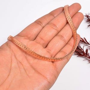 Puede incluir: Un collar de cadena de serpiente plana de oro rosa. La cadena se sostiene en una mano sobre un fondo blanco. El collar tiene un patrón tejido y es de un color metálico cálido.