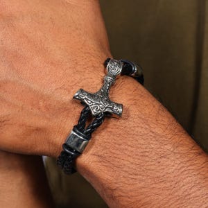 Puede incluir: Un brazalete con un colgante de martillo de Thor plateado y una banda de cuero trenzado negro. El martillo presenta detalles intrincados. El brazalete se asegura con un cierre plateado.
