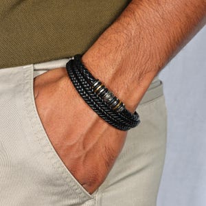 Könnte beinhalten: Schwarzes geflochtenes Lederarmband mit silber- und goldfarbenen Metallakzenten. Das Armband hat mehrere Stränge aus geflochtenem Leder und dekorativen Metallperlen. Ein stilvolles Accessoire.