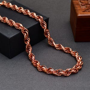Puede incluir: Un collar de cadena color cobre con un diseño grueso de cuerda trenzada compuesto por eslabones circulares entrelazados. La superficie metálica tiene un acabado pulido y reflectante que capta la luz, creando un patrón continuo y texturizado.