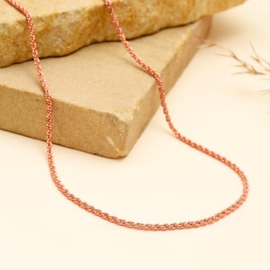 May include: A delicate rose gold chain necklace. The necklace is displayed on a neutral background with a textured stone. The chain has a woven design and is a warm, copper color.