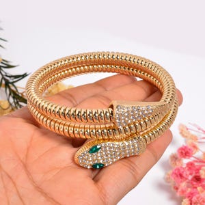 Peut inclure: Bracelet serpent doré avec un design enroulé. La tête et la queue du serpent sont ornées de strass transparents et d'accents verts. Le bracelet est enroulé autour d'une main, mettant en valeur ses détails.