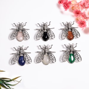 Puede incluir: Seis broches decorativos con forma de abeja con alas y cuerpos de metal plateado. Cada abeja presenta una gema diferente: cuarzo rosa, ónix negro, piedra solar, lapislázuli, cuarzo transparente y malaquita. Los broches se muestran sobre una superficie blanca.
