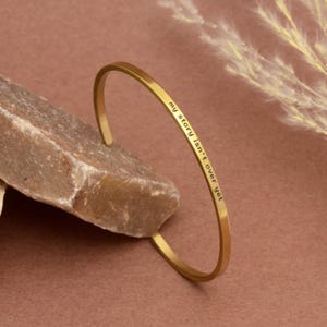 Könnte beinhalten: Ein goldfarbenes Armreif-Armband mit der Aufschrift "my story isn't over yet". Das Armband liegt auf einem hellbraunen Stein vor einem weichen braunen Hintergrund. Das Armband hat ein schlichtes, elegantes Design.