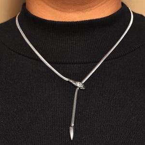 May include: A silver-toned necklace with a snake-shaped pendant and a dangling teardrop-shaped charm. The necklace is worn against a black top, highlighting the jewelry's design and color.