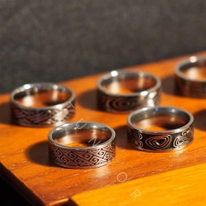 Może przedstawiać: Kolekcja srebrnych pierścionków z różnymi wytrawionymi wzorami, prezentowana na drewnianej powierzchni. Pierścionki mają geometryczne i abstrakcyjne wzory, prezentujące różnorodność faktur i stylów. Pierścionki są prezentowane w dobrze oświetlonym otoczeniu, podkreślając ich metaliczny połysk.