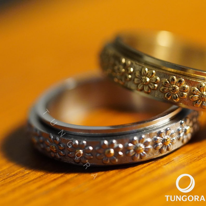 Close-up of a silver daisy flower spinner ring with intricate floral engravings and golden centers, photographed on a warm wooden surface. Elegant boho jewelry, mindfulness fidget ring, stress relief spinner band for women, daisy flower design, nature-inspired gift, floral meditation jewelry.