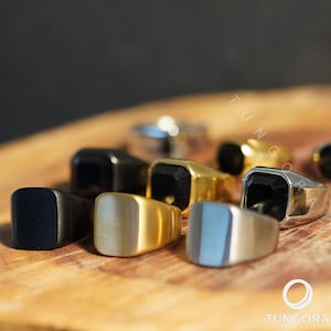 Op de afbeelding: Een verzameling zegelringen in verschillende kleuren en afwerkingen, waaronder zwart, goud en zilver. Sommige ringen hebben een zwarte edelsteen. De ringen worden gepresenteerd op een houten oppervlak. De merknaam "TUNGORA" is zichtbaar.