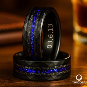 May include: Two black tungsten rings with a faceted design. Each ring features a blue inlay and a decorative pattern. One ring is engraved with the date "03.6.13". The rings are displayed on a wooden surface.