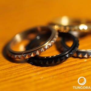 Puede incluir: Primer plano de varios anillos con un diseño texturizado y puntiagudo en los bordes. Los anillos son de varios colores, incluyendo plata, negro y oro. Los anillos están sobre una superficie de madera.