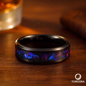 Könnte beinhalten: Ein schwarzer Ring mit einem Band aus blauem und violettem schillerndem Material. Der Ring liegt auf einer Holzoberfläche. Der Markenname "Tungora" ist unten rechts sichtbar.