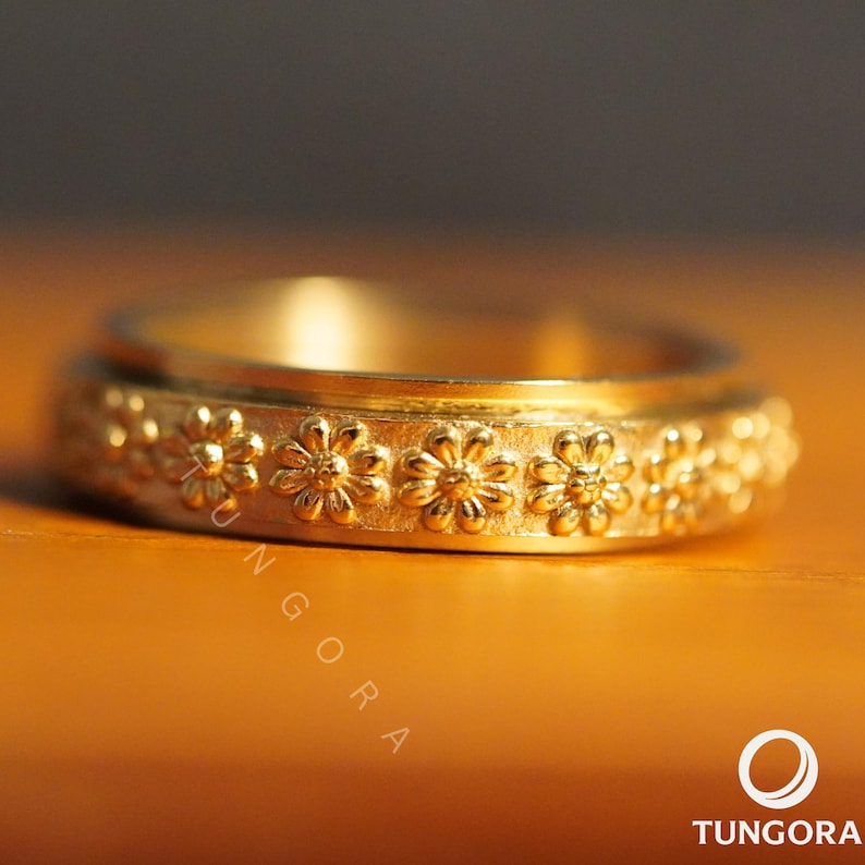 Op de afbeelding: Een goudkleurige ring met een bloemenontwerp. De ring heeft een band met reli&euml;fbloemen rondom. De achtergrond heeft een warme, oranje tint. De merknaam "TUNGORA" is rechtsonder zichtbaar.