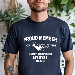 May include: Navy blue t-shirt with the text "PROUD MEMBER OF THE JUST RESTING MY EYES CLUB" in white. The graphic shows a person reclining in a chair. The shirt is made of a soft material.